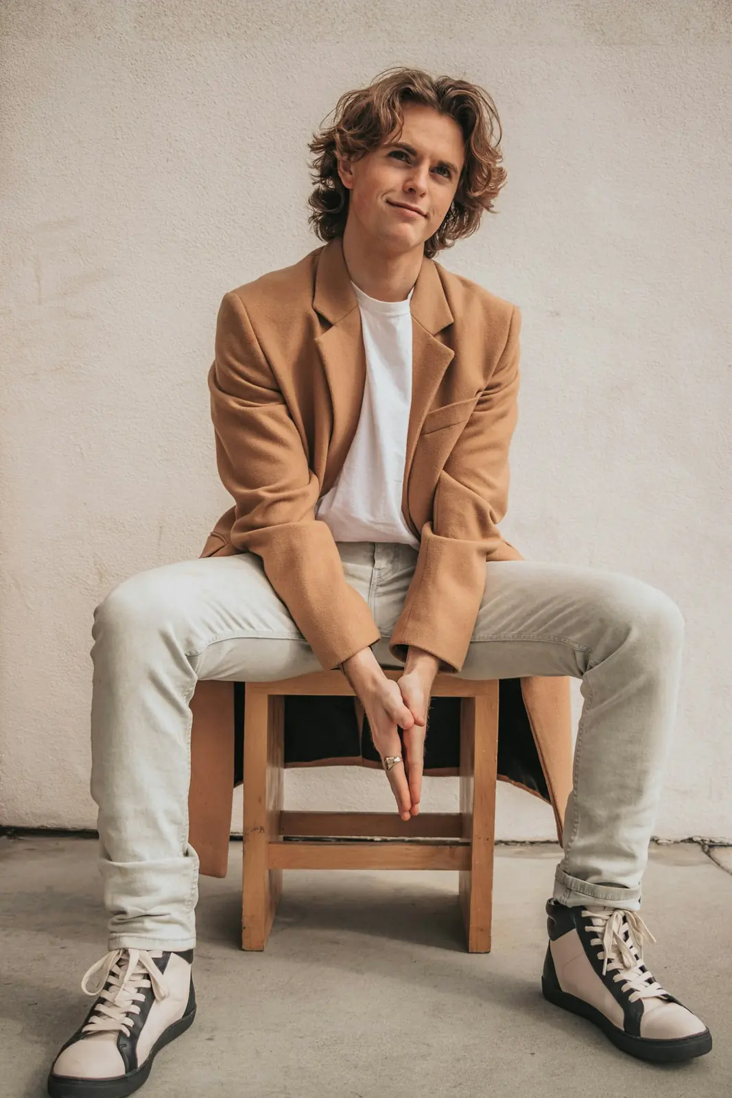 A man seated on a stool in a camel blazer and white tee